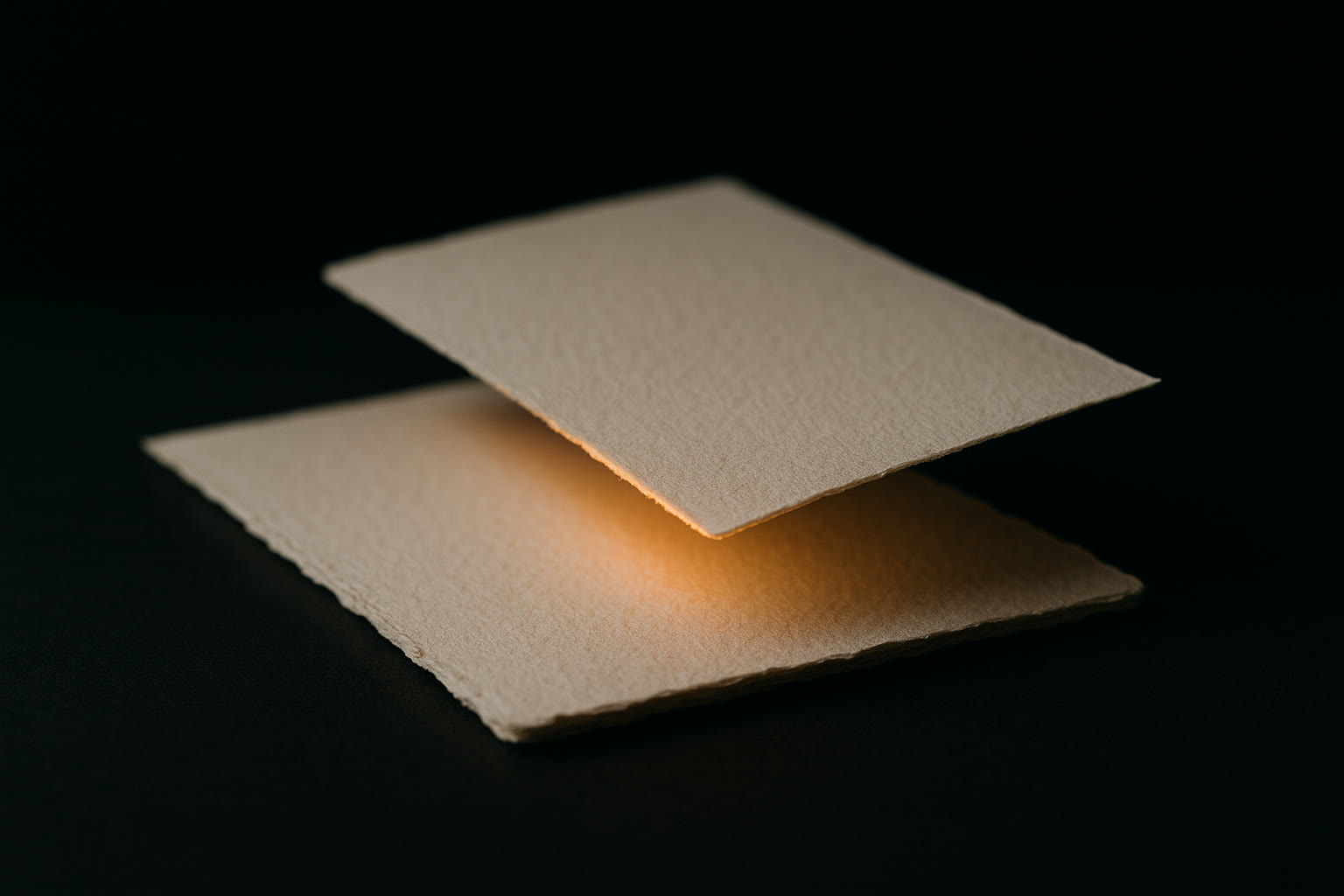 Two sheets of handmade paper on a dark table, one sliding over the other, lit from between by a warm amber glow.